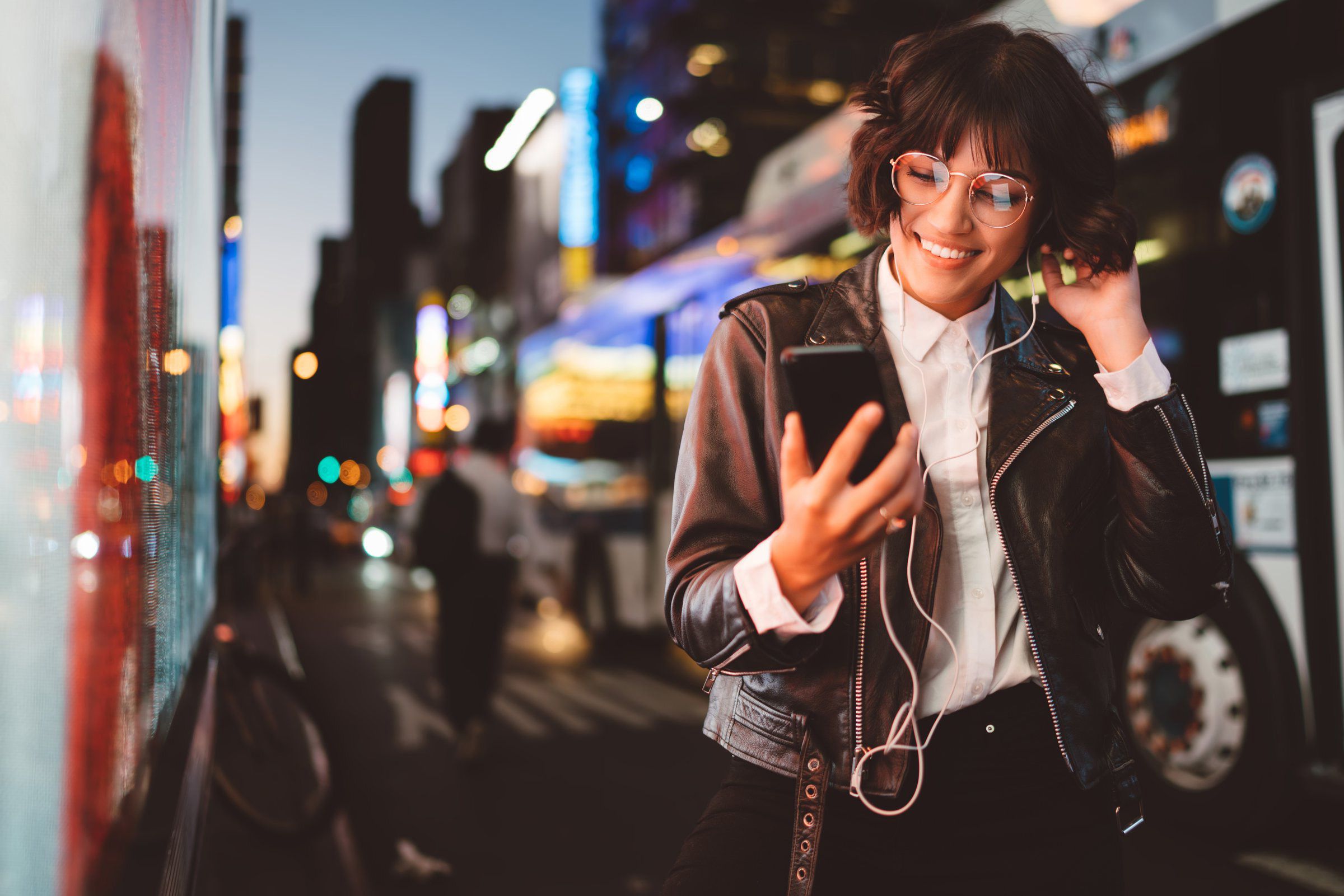 Cheerful pretty young woman in cool eyeglasses and trendy wear walking on metropolis street with night lights enjoying songs from playlist in earphones and reading sms with good news on smartphone